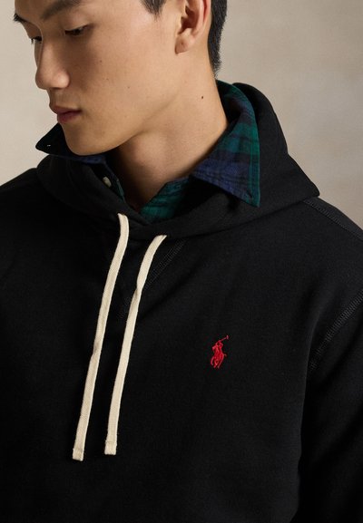 Man wearing black hoodie with white drawstrings and red embroidered logo, over a green and navy checkered collared shirt.