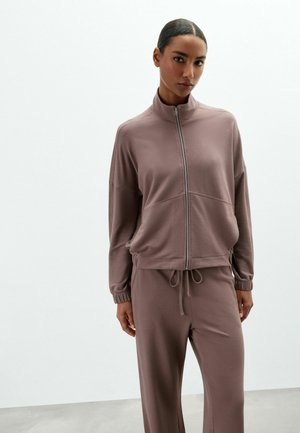 Frau mit nach hinten gegelten Haaren, die eine mauve Zip-Up-Jacke und passende Hose trägt und vor einem schlichten weißen Hintergrund steht.