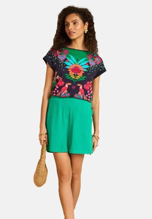 Nainen, joka pukeutunut värikkääseen kukka- ja lintukuviolliseen yläosaan sekä vihreisiin korkeavyötäröisiin shortseihin, pitäen vasemmassa kädessään punottua käsilaukkua.