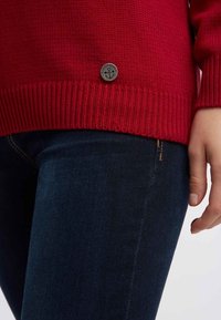 Roter Strickpullover mit geripptem Saum, der ein kleines rundes Metallschlossdetail aufweist. Kombiniert mit dunkelblauen Jeans, Nahaufnahme des Oberkörpers.