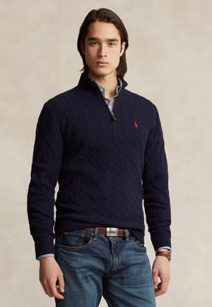 Jeune homme aux cheveux foncés portant un pull en maille torsadée bleu marine, une chemise rayée, un jean bleu, une ceinture marron et une montre marron, debout devant un fond uni.