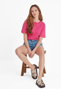 Bright pink cotton T-shirt with short sleeves, paired with high-waisted denim shorts. Model wearing grey slide sandals with buckles.