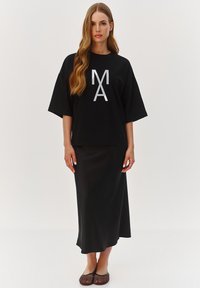 Przodowy czarny t-shirt w rozmiarze oversize z białą grafiką, w zestawieniu z czarną satynową spódnicą midi i brązowymi perforowanymi płaskimi butami. Gładkie tekstury w całym stroju.