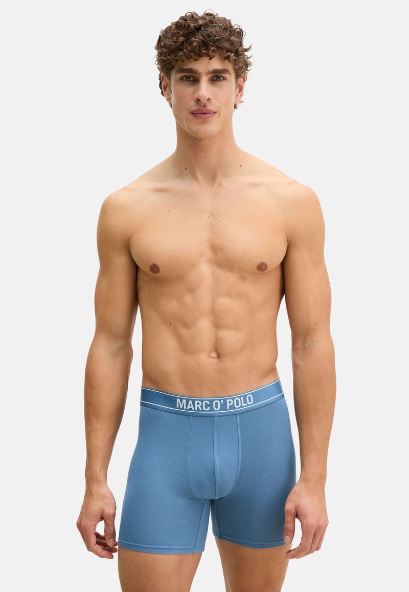 Homme athlétique aux cheveux bouclés portant un boxer Marc O'Polo bleu, debout devant un fond gris clair uni.
