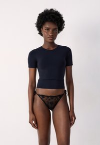 Haut ajusté à manches courtes bleu marine associé à un string en dentelle noire avec des motifs floraux et des liens sur les côtés. Texture lisse sur l'ensemble.