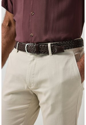 Primer plano de una persona con pantalones beige, cinturón de cuero trenzado marrón oscuro y camisa granate metida por dentro.