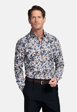 Erwachsener Mann trägt ein langärmeliges, geblümtes Hemd in Blau-, Braun- und Weißtönen, mit schwarzen Hosen und einem braunen Gürtel.