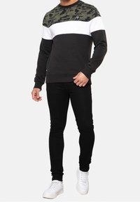 Zwarte sweatshirt met een groen camouflagepatroon aan de bovenkant, een witte horizontale streep, geribbelde boorden en zoom, gecombineerd met zwarte skinny jeans en witte sneakers.