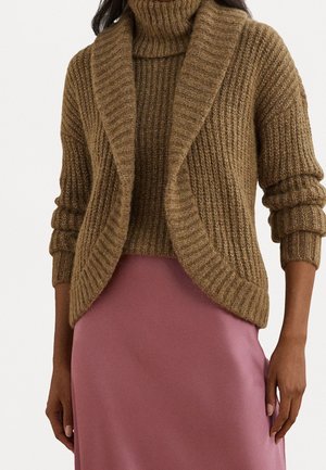 Femme portant un pull à col roulé marron en maille côtelée avec un cardigan assorti et une jupe rose soyeuse, debout, les bras détendus.