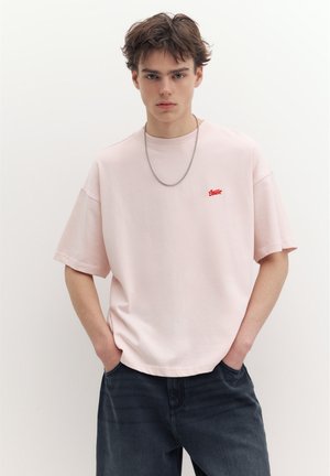 Ung mand med kort brunt hår iført en lyserød oversized T-shirt med et lille rødt logo og mørke jeans, hænderne i lommerne, neutralt baggrund.