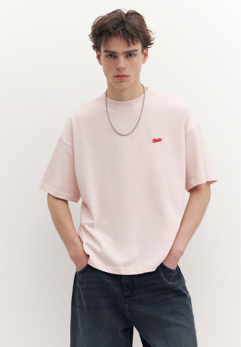 Jeune homme aux cheveux bruns courts portant un t-shirt ample rose clair avec un petit logo rouge et un jean foncé, mains dans les poches, fond neutre.