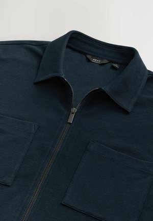 Dark navy zippered shirt featuring a textured fabric, pointed collar, and two front patch pockets with square openings.