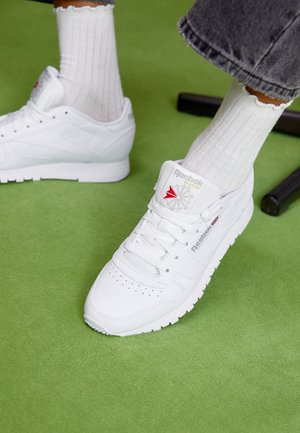 Baskets blanches Reebok Classic portées avec des chaussettes blanches côtelées et un pantalon gris, debout sur un tapis vert près des pieds noirs d'une chaise.