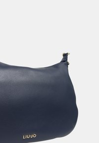 Marineblauwe leren schoudertas met een gladde textuur, gebogen vorm, gouden hardware accenten en een merkembleem aan de onderkant.