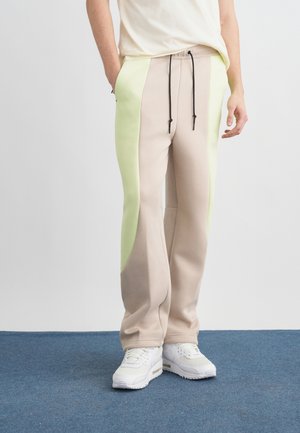 Persona che indossa pantaloni sportivi a blocchi di colore beige e verde chiaro con coulisse nere e sneakers bianche, in piedi su un tappeto blu.