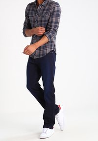 Rutig skjorta i blått, grått och vitt med två bröstfickor; mörka jeans; vita sneakers med röda detaljer; avslappnad outfit.