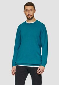 Türkisfarbener Rundhals-Pullover aus glattem Material, mit langen Ärmeln, einem weißen Innensaum und gerippten Bündchen. Kombiniert mit schwarzen Hosen.