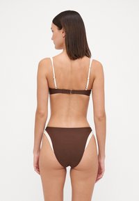 Brūns bikini komplekts ar regulējamiem baltiem siksnām, aizdari aizmugurē un augsto vidukli ar sānu izgriezumiem. Vienmērīga tekstūra.