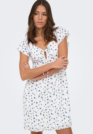 Femme portant une robe blanche avec de petits motifs floraux bleus, des manches courtes volantées et une découpe nouée à l'avant, debout, les bras croisés.