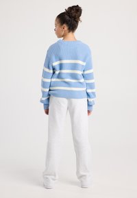 Young person standing facing away, wearing a blue and white striped sweater, white pants, and white shoes against a plain background.