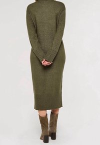 Femme debout, les mains jointes dans le dos, portant une robe midi en maille vert olive et des bottes à talons assorties en daim.