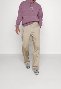 Felpa con cappuccio viola con una toppa logata, pantaloni beige e sneakers multicolori, dotate di una suola imbottita e lacci.