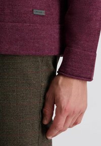 Geweven bordeauxrode trui met een label, gecombineerd met bruine geruite broek. De focus ligt op de details van de mouw en hand, waarbij de stof en pasvorm worden getoond.
