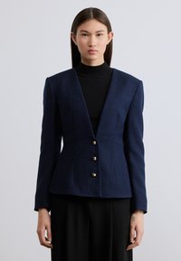 Μπλε ναυτικό σακάκι από υφασμάτινο ύφασμα με υφή, με βαθύ ντεκολτέ σε σχήμα V, δομημένους ώμους και τρία κούμπωμα με χρυσά κουμπιά μπροστά.