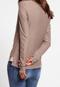 Top de manga larga color beige con un ligero escote en V, dobladillo redondeado y aberturas laterales, llevado sobre jeans azules. Textura de punto suave, estilo ajustado.