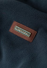Tejido de forro polar azul marino con un parche de cuero marrón que muestra el logo "SUPERDRY®" en letras doradas, sobre un fondo texturizado.