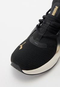 Sort atletisk sneakers med en struktureret mesh-overdel, guld Puma-logo og sorte snørebånd. Sålen er hvid med en let bue ved tæerne.