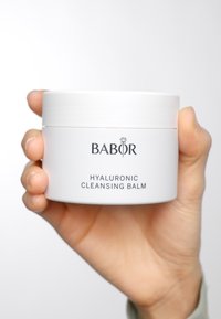 Witte ronde container met een gladde oppervlakte, gelabeld "BABOR Hyaluronic Cleansing Balm" in zwarte lettertype. Vastgehouden in een hand, minimalistisch ontwerp.