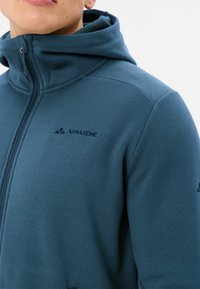 Giacca in pile teal con cappuccio, dotata di zip integrale e dettagli con logo. Testura liscia e vestibilità atletica con cuciture decorative sulle spalle.