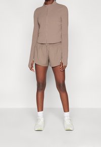 Top lungo a maniche lunghe con zip di colore marrone abbinato a pantaloni corti coordinati. Tessuto morbido, design aderente e vita elastica. Le scarpe sono grigie chiare con dettagli colorati.
