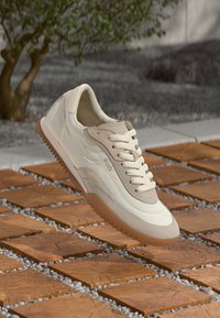 Scarpa sportiva beige con tomaia in tessuto morbido, lacci anteriori e suola in gomma. Presenta linee di design curve e accenti tonali.