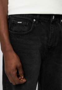 Pantalones vaqueros de mezclilla negra con una textura sutil, que presentan un bolsillo delantero, herrajes plateados y una pequeña etiqueta de la marca en la cinturilla.