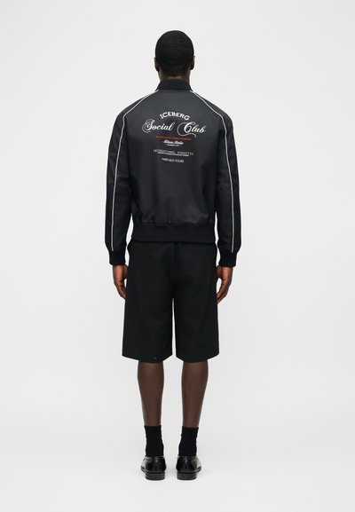 Άνδρας που φοράει μαύρο μπουφάν bomber με λευκή διακόσμηση και λευκό κείμενο "Iceberg Social Club" στην πλάτη, συνδυασμένο με φαρδιά μαύρα σορτς και μαύρα παπούτσια.