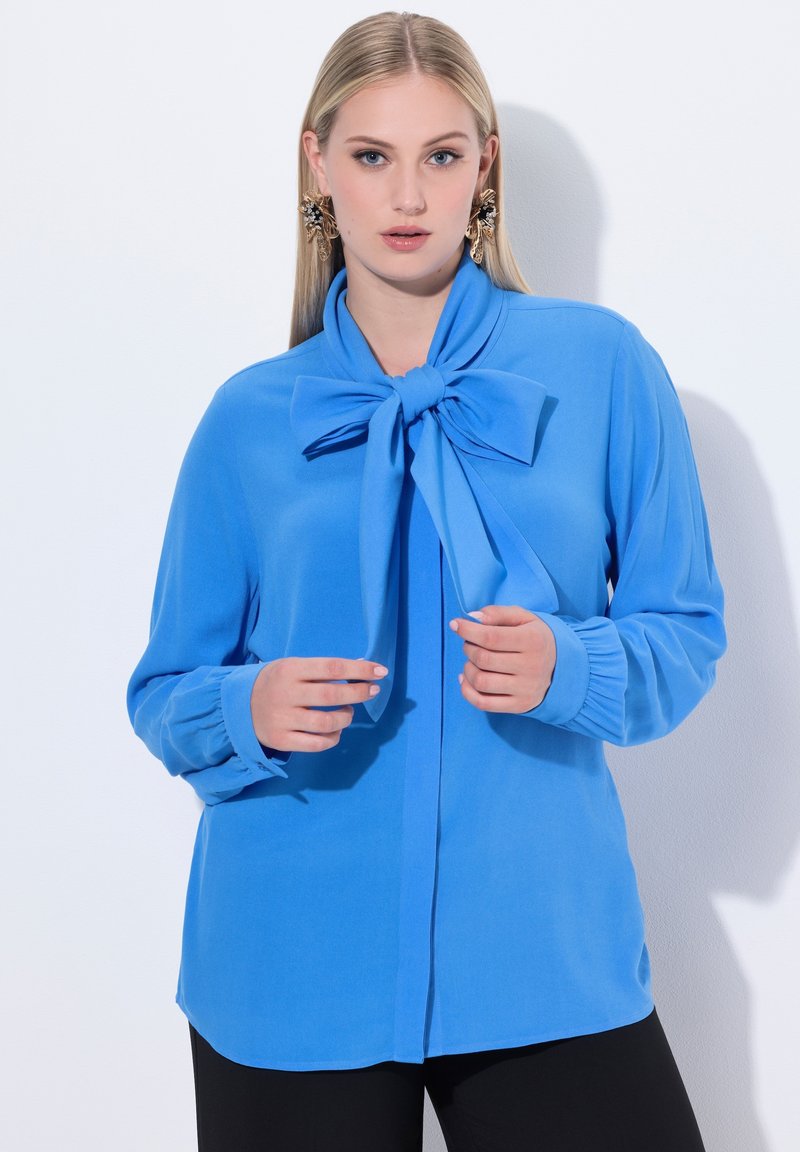 Blusa azzurro chiaro con un grande fiocco al collo, maniche lunghe e polsini arricciati. Tessuto morbido e liscio con una vestibilità su misura.