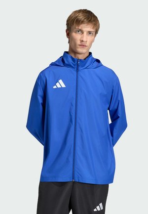 Giovane uomo che indossa una giacca Adidas blu con zip e cappuccio e pantaloni Adidas neri, in piedi davanti a uno sfondo chiaro e uniforme.