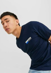 Tumši zils kokvilnas T-krekls ar baltu un zilu logo plāksteri uz krūtīm. Īsas piedurknes, apaļa kakla izgriezne un maiga tekstūra.