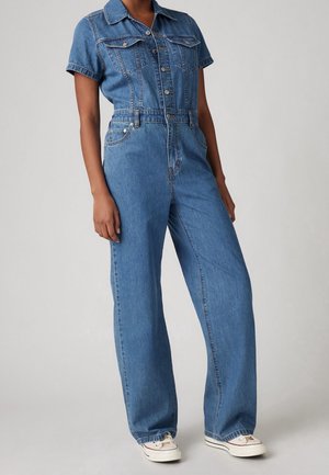 Person iført en blå denim kortærmet jumpsuit med knapper og hvide sneakers mod en ensfarvet lys baggrund.