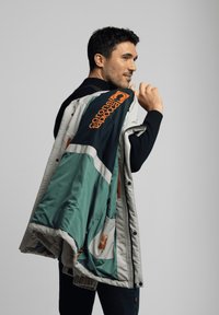 Hombre sosteniendo una chaqueta verde y gris con el logo naranja "Capronel Abbiocca" sobre su hombro, mirando hacia la derecha sobre un fondo blanco.