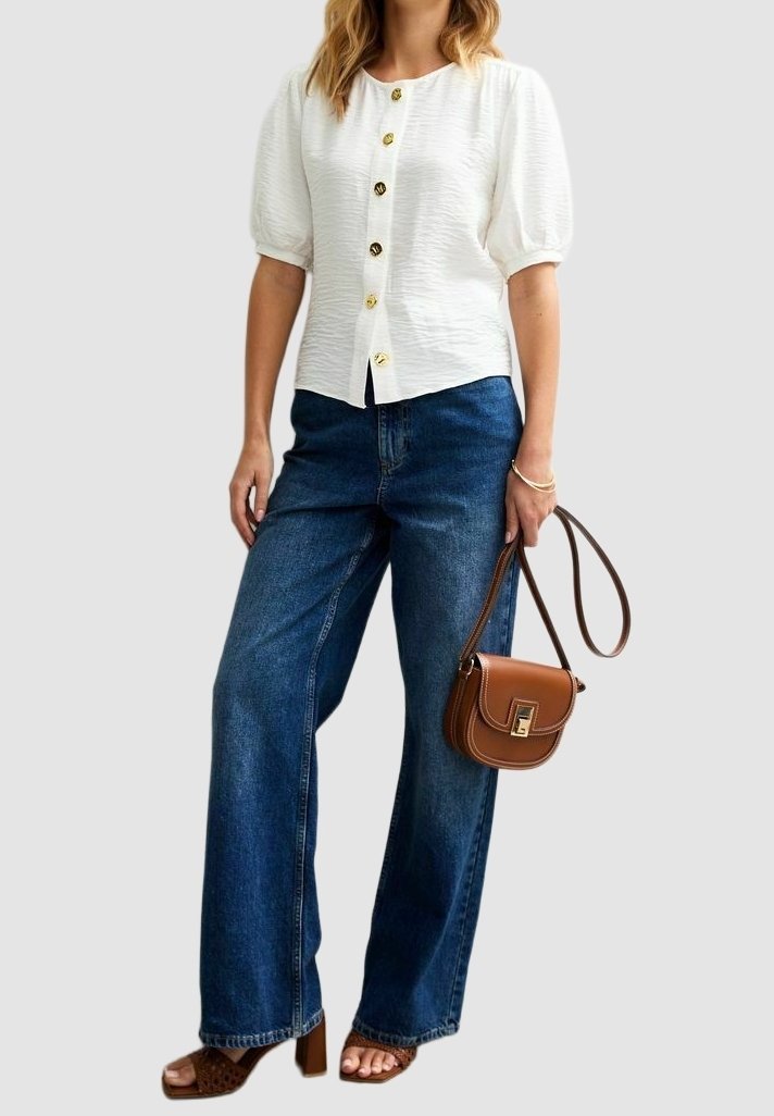 Femme portant un chemisier blanc à manches courtes boutonné, un jean bleu à jambes larges, des sandales marron à talons et tenant un petit sac à main en cuir marron.