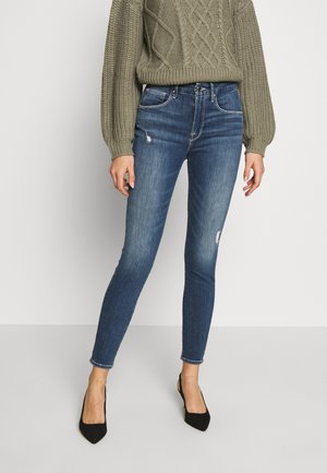 Frau trägt blaue Skinny-Jeans mit leichter Distressed-Optik, kombiniert mit einem olivgrünen Strickpullover und schwarzen spitzen Absätzen.