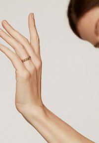 Mano levantada con los dedos extendidos, portando un anillo de oro con pequeños diamantes en el dedo anular, fondo claro y liso.