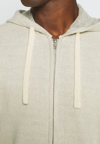 Felpa con cappuccio grigia chiara a maglia con zip completa, cappuccio regolabile e texture morbida. Presenta una zip argentata e stringhe color crema.