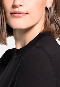 Suéter negro de cuello alto con textura suave, presenta un cuello alto y costuras lisas. Se puede ver el corte de pelo bob del modelo y unos sutiles pendientes de aro plateados.