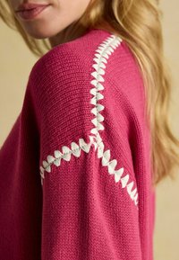 Pulover tricotat fucsia, cu un finisaj texturat, având o cusătură albă în zigzag pe linia umărului. Material moale, cu greutate medie.