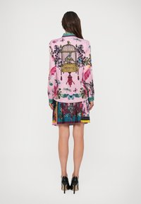 Vrouw staat met haar rug naar ons toe en draagt een roze blouse en een geplisseerde rok met prints van vogelkooien, bloemen, vlinders en libellen, gecombineerd met zwarte hakken.