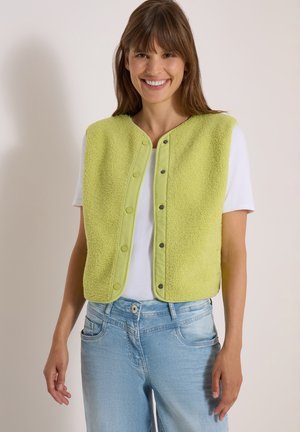Kvinde iført en lysegrøn fleecevest med knapper over en hvid t-shirt og lyseblå jeans, smilende og stående mod en ensfarvet baggrund.